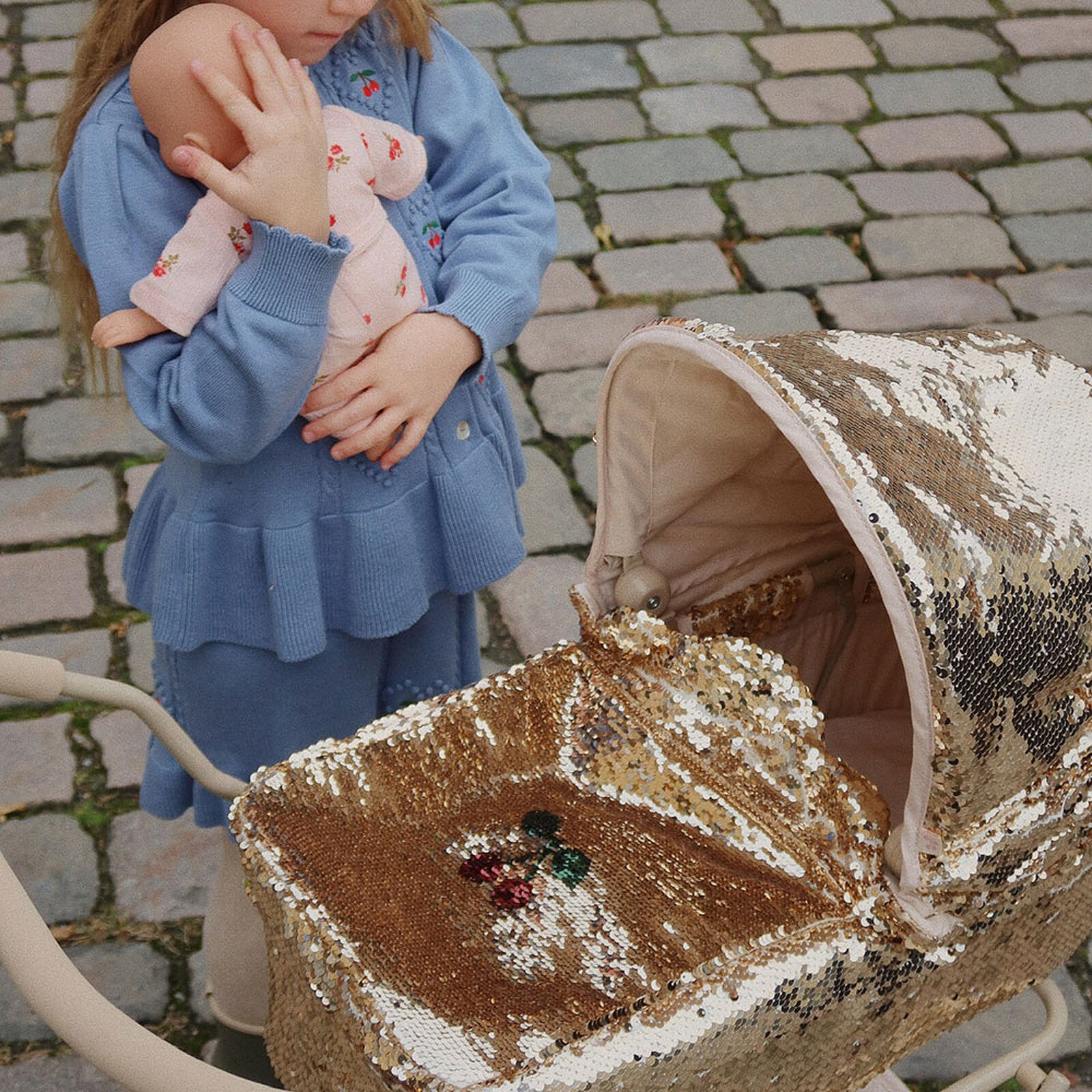 Petite fille tenant son poupon à côté d’une poussette dorée pour poupée, jouet élégant et scintillant.