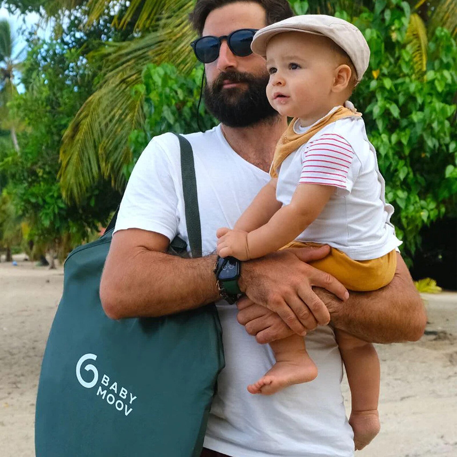 Papa portant son bébé sur la plage avec le sac de plage Babymoov et le set de jouets Swim Essentials – moment de détente en famille au bord de la mer
